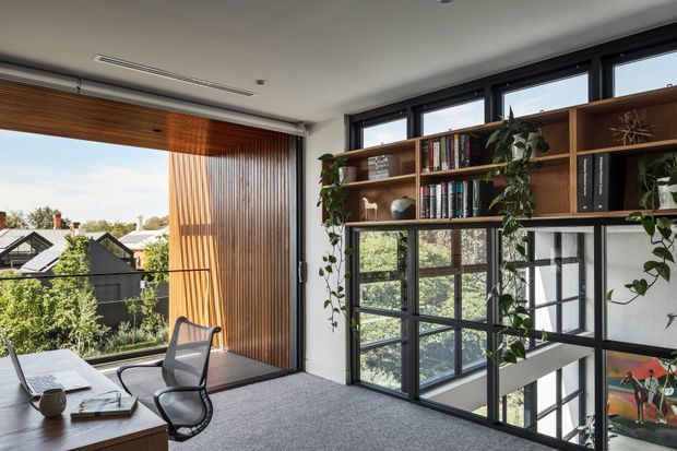 A bright contemporary home office with a large desk, ergonomic chair, dark-framed floor-to-ceiling glazing, and an internal black-framed glass balustrade overlooking a lower level. A timber venetian blind and vertical timber-lined wall add warmth, while built-in open shelving displays books, plants, and decorative objects. The room has a grey carpet, white ceiling and walls, and expansive leafy suburban views.