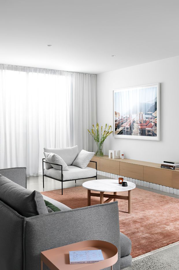 Bright, refined contemporary living room with floor-to-ceiling sheer curtains, pale walls, a grey upholstered sofa and armchair, a round white coffee table, a long timber media console, and a soft blush-toned rug. A framed artwork provides a subtle focal point, while the layered neutral palette creates an airy, calm atmosphere.