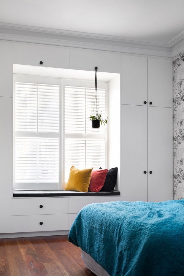 Bright, compact bedroom with extensive custom white built-in cabinetry surrounding a window seat. White plantation shutters fill the window, with a hanging plant and three colourful cushions on the dark upholstered bench. A vivid turquoise quilt covers the bed in the foreground, complemented by warm timber flooring, ornate ceiling cornices, and patterned botanical wallpaper on the right wall.
