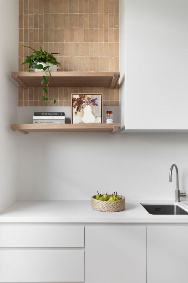 A bright, minimalist kitchen vignette with white flat-panel cabinetry, a white benchtop, and a stainless-steel sink and gooseneck tap. Two floating timber shelves display a potted plant, books, artwork, and small decor against a vertical tan ceramic tile feature wall. The overall palette is white, warm timber, and muted beige with clean contemporary detailing.