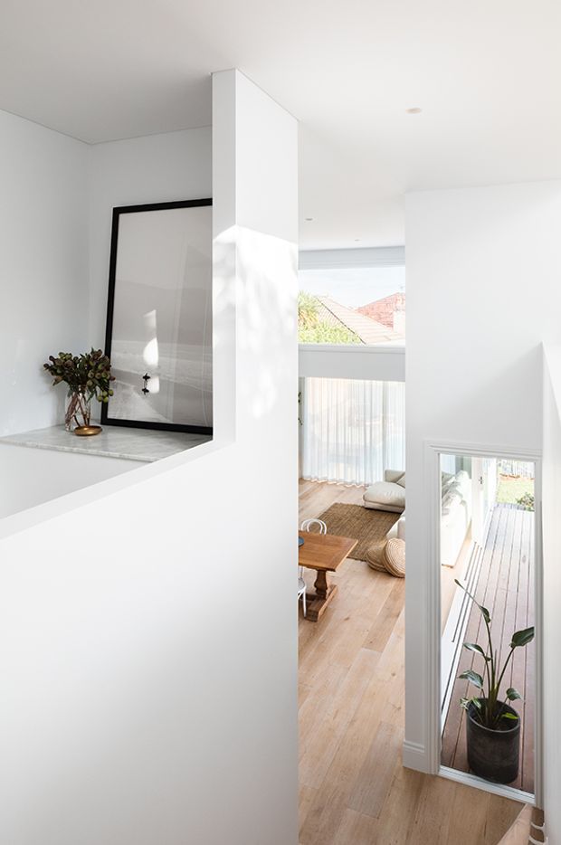 Bright minimalist interior view from a stair or circulation area toward an open-plan living space and glazed exterior doorway. White walls and ceiling, a large black-framed artwork, pale timber flooring, a stone ledge, and abundant natural light define the scene. The view includes a tall void or open connection between levels, with a deck visible through the glazed door.