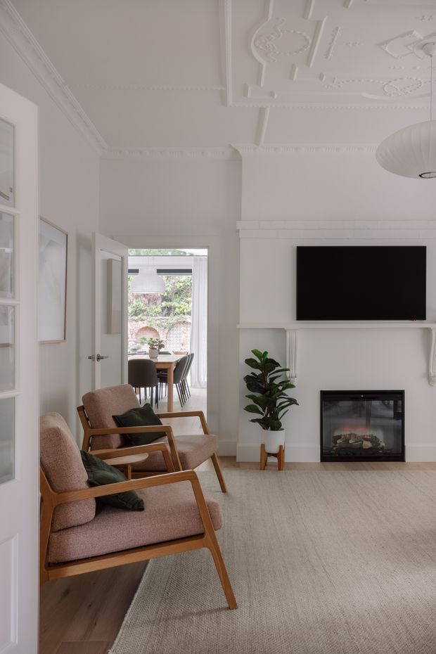 Bright, refined living room with ornate white decorative ceiling mouldings, a classic fireplace mantel with a wall-mounted television, and two timber-framed upholstered lounge chairs. A large neutral rug covers much of the timber floor, while a doorway opens to a dining area and garden beyond. The room combines traditional architectural detailing with restrained contemporary furnishings.