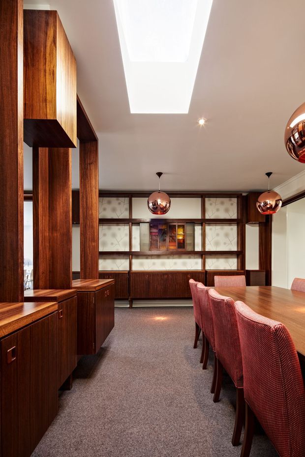 A richly detailed dining room with extensive dark timber built-in cabinetry and wall panelling, including a geometric open partition on the left and a full-height display/storage wall at the back. A long timber dining table is surrounded by upholstered reddish-pink chairs. Three reflective copper-toned spherical pendant lights hang above the table, while a large rectangular skylight brightens the ceiling. The room has carpeted flooring, warm neutral walls, and a refined retro-inspired character.