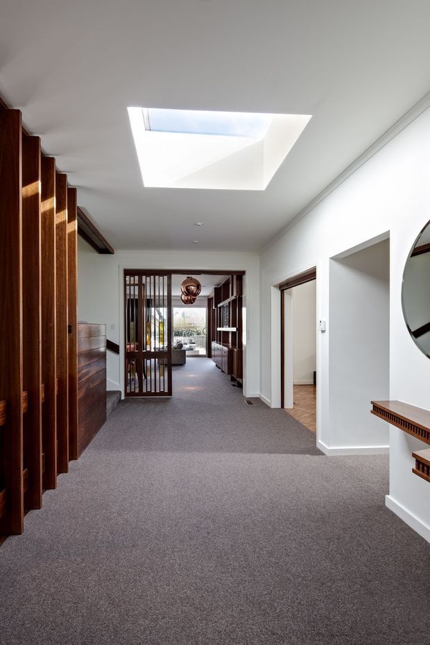 A bright residential hallway or landing with grey carpet, white walls and ceiling, and a large rectangular skylight. Dark timber vertical slatted screening and built-in joinery line the left side, while timber-framed doorways and shelving lead toward a living area at the rear. A round wall mirror and narrow timber console are visible on the right. The space has a refined mid-century-influenced character with strong natural light and warm timber detailing.