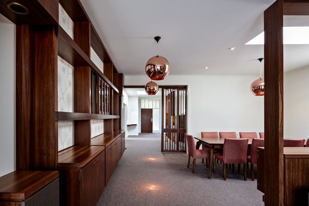 A refined open-plan dining and living area with extensive dark timber built-in cabinetry and shelving, decorative vertical timber screens, a long dining table with upholstered pink chairs, and three copper pendant lights. The room has white walls and ceiling, grey carpet, and a restrained mid-century-inspired interior with strong timber detailing and warm metallic accents.