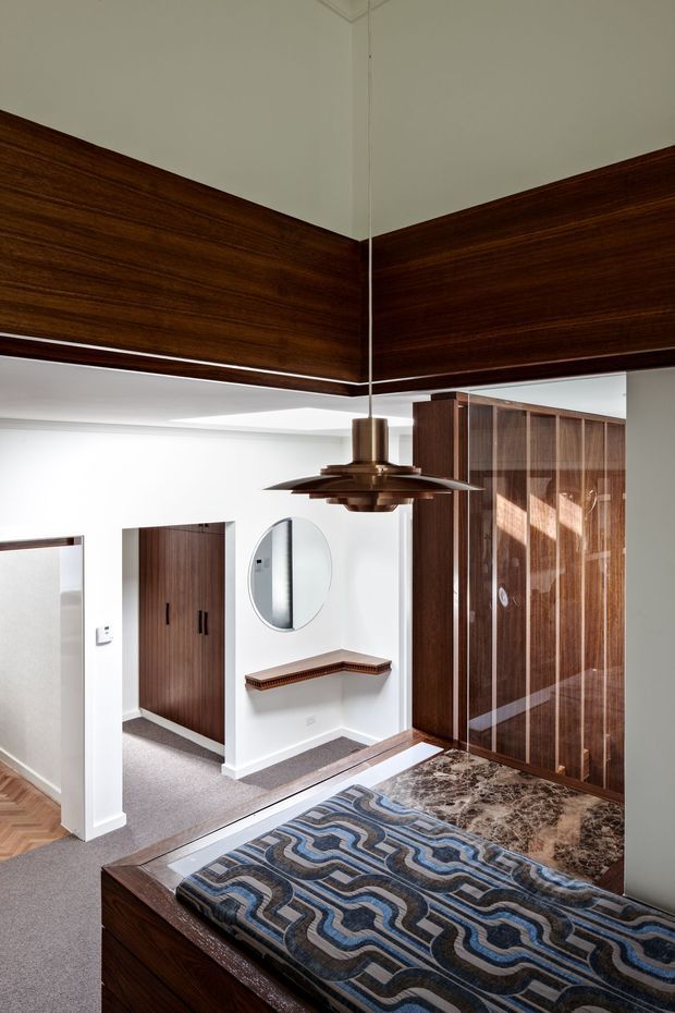 A richly detailed bedroom interior with dark wood wall panelling and built-in wardrobes, a patterned upholstered bed, marble bedside surface, and a sculptural suspended pendant light. White walls and ceiling provide contrast, while a hallway and circular mirror are visible beyond the room. The composition has strong mid-century character with polished timber joinery and geometric textiles.