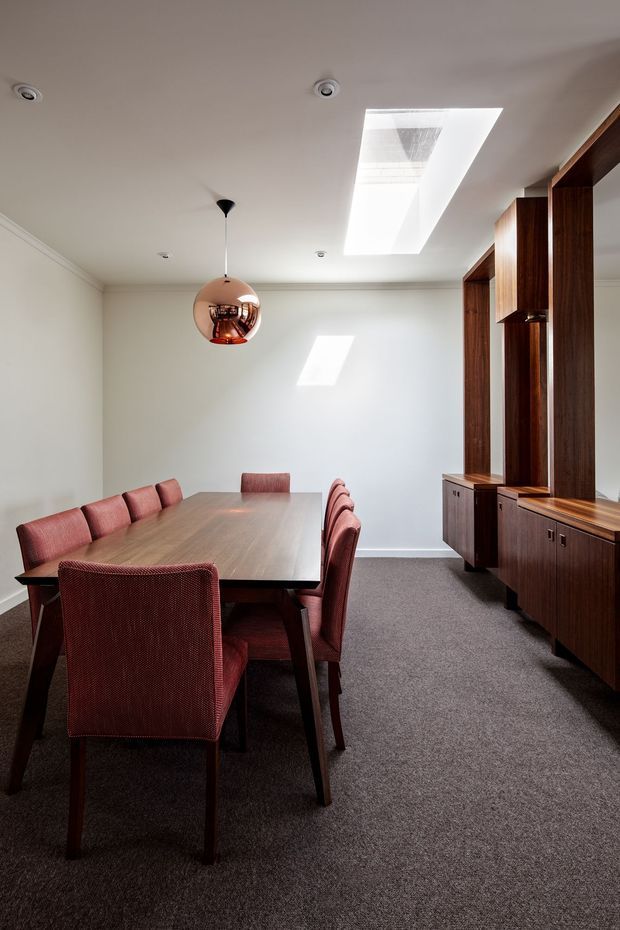 A refined dining room with a long timber dining table surrounded by upholstered burgundy-red chairs. The room has white walls and ceiling, grey carpet, a large skylight, recessed ceiling lights, a copper spherical pendant, and extensive dark timber built-in cabinetry with open display niches along the right wall.