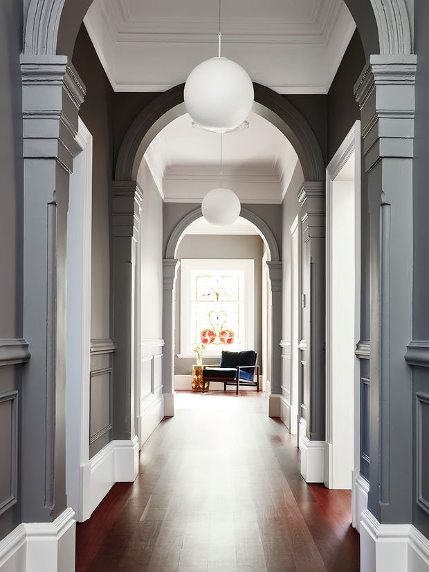 A grand, symmetrical interior hallway with repeated arched openings, dark grey decorative wall panelling, white architectural trim, coffered ceilings, polished dark timber flooring, and spherical pendant lights. A bright stained-glass window and a small armchair are visible at the far end.