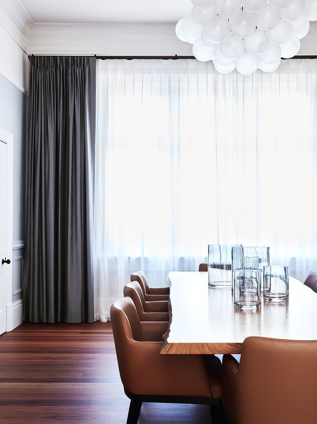 A bright, refined dining room with a long contemporary dining table and warm brown leather upholstered chairs. Floor-to-ceiling sheer curtains are paired with dark charcoal drapes, while white decorative wall panelling and moulding frame the room. A sculptural cluster pendant light hangs overhead, and dark timber flooring adds warmth and contrast.