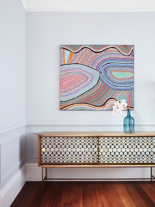 A refined interior vignette featuring a long timber sideboard with decorative circular-patterned doors and slender metal legs, set against pale blue-grey wall panelling with white moulded trim. A colourful abstract artwork hangs above, accompanied by a blue glass vase and white flowers. Dark timber flooring and restrained styling create a polished, layered look.