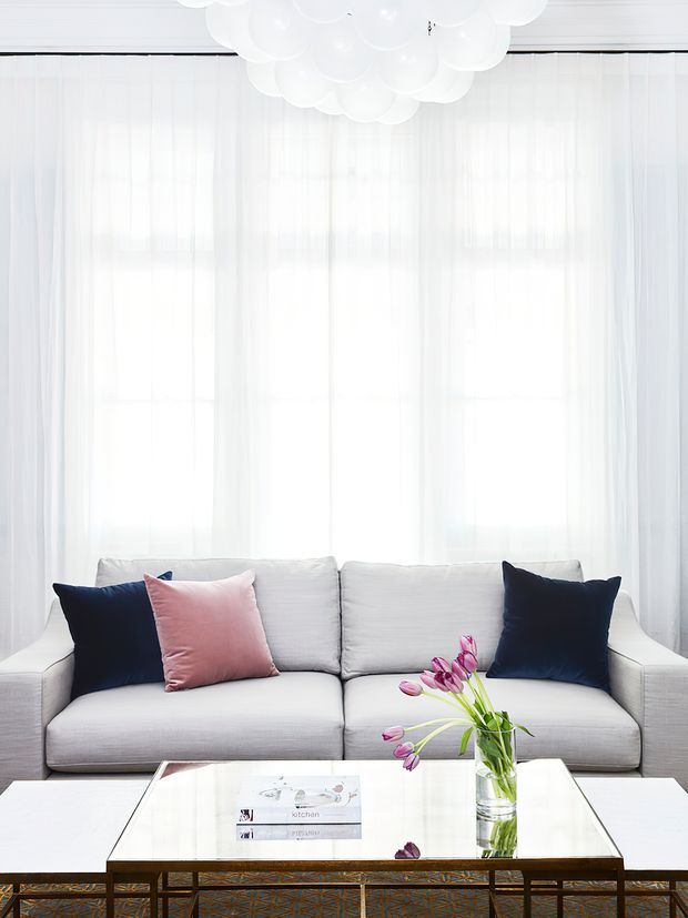 Bright, airy living room with a pale upholstered sofa positioned in front of full-height sheer white curtains. Navy and blush cushions add contrast, while a reflective rectangular coffee table with a dark metal base holds a glass vase of pink tulips. A sculptural cluster-style white pendant light hangs overhead.