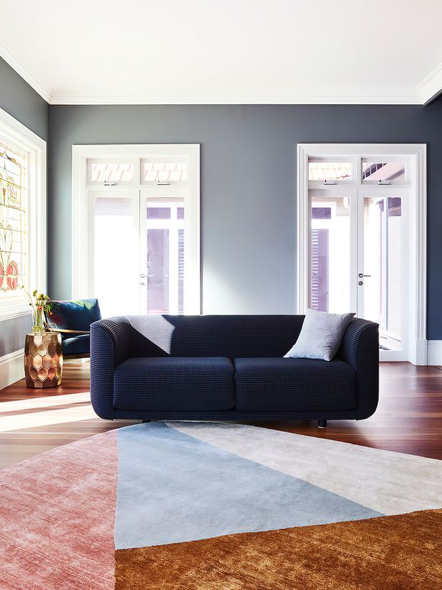 Bright, refined living room with dark blue-grey walls, white trim, tall glazed doors and windows, polished timber flooring, a navy upholstered sofa, and a large geometric area rug in blush pink, pale blue, cream, and ochre. A stained-glass window and decorative side table add character.