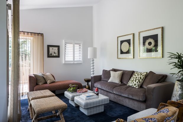 A bright, comfortably furnished living room with a large taupe sectional sofa, a burgundy chaise lounge, woven upholstered ottomans, and a dark blue area rug. The room features white walls, plantation shutters, sheer curtains, framed botanical-style artwork, a sculptural table lamp, and indoor greenery.