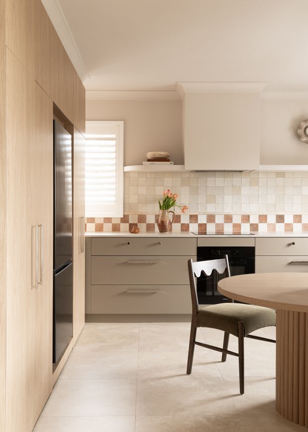 Warm, refined contemporary kitchen with light timber full-height cabinetry, taupe-grey drawers, a white rangehood and tiled splashback. The splashback combines cream square tiles with a distinctive terracotta-and-cream checkerboard band. A round fluted timber dining table and upholstered dark wood chair occupy the foreground, while integrated appliances and soft natural light complete the space.