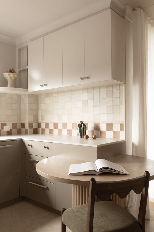 A warm, compact kitchen-dining area with cream cabinetry, a white benchtop, tiled backsplash, and a small round timber dining table beside a bright curtained window. The cabinetry includes upper wall cupboards and lower drawers, while the backsplash combines pale square tiles with a terracotta-and-cream checkerboard accent band. A vase and open book add domestic detail.