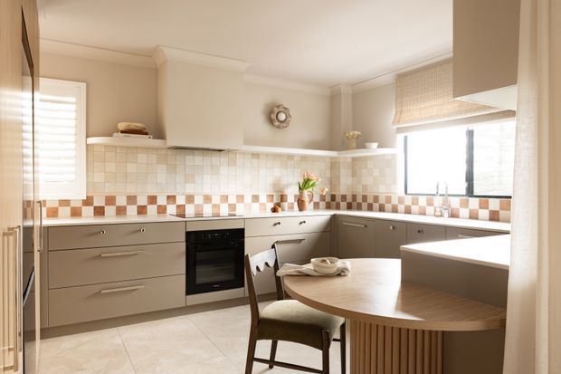 A warm, bright kitchen with taupe-grey cabinetry, integrated oven, tiled backsplash, pale tiled flooring, and a rounded dining island with vertical timber detailing. Cream walls, plantation shutters, woven roller blinds, and natural wood accents create a refined traditional-contemporary interior.