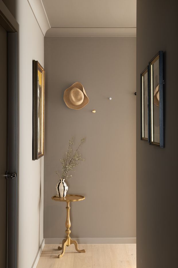 A narrow, warmly lit interior hallway or entry passage with taupe walls, a high decorative ceiling trim, pale timber flooring, framed artwork, and a small round wood pedestal table displaying a monochrome vase with delicate branches. Mirrors or reflective framed panels line the sides, creating depth. The styling is refined and understated with warm neutral tones and classic decorative details.