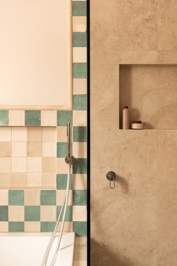 A close-up of a warmly styled bathroom featuring a bathtub or shower area with a retro checkerboard arrangement of cream and muted sage-green ceramic tiles, alongside a contemporary beige stone-look tiled wall. A recessed wall niche holds toiletry bottles, and chrome shower controls and a handheld shower hose are visible. The palette is warm, earthy, and softly vintage-inspired.