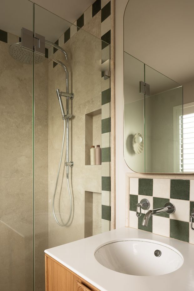 A compact, warmly lit bathroom with a clear glass walk-in shower, large chrome rain shower and handheld fixture, beige stone-look wall tiles, and recessed shower niches. A white inset basin sits in a pale benchtop above a timber vanity. The splashback features a checkerboard pattern of dark green and off-white square tiles, echoed in a vertical shower border. A large rounded-corner mirror and shuttered window reflect and brighten the space.