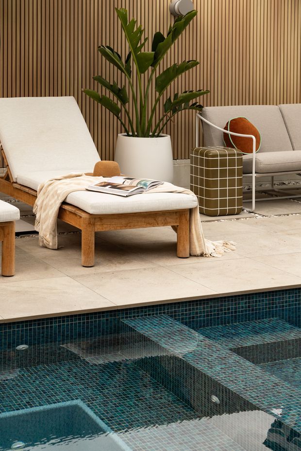 A refined poolside lounge area with a stepped swimming pool in the foreground. The terrace is paved in large pale stone slabs and furnished with a timber-framed chaise lounge, matching upholstered ottoman, and contemporary sofa. A tall potted tropical plant stands against a vertical timber slat wall, while a rounded grid-patterned side table and draped neutral throw add texture. The pool is finished in small dark teal mosaic tiles with integrated submerged seating or steps. The overall look is warm, contemporary, and resort-like.