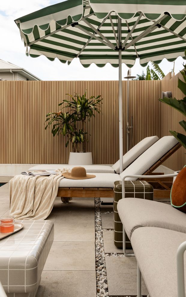 A stylish outdoor lounge and poolside-style entertaining area enclosed by a vertical timber privacy fence. The space features a large green-and-white striped scalloped-edge umbrella, upholstered sun loungers, a cushioned outdoor sofa and ottoman, potted tropical greenery, light stone paving, and a decorative pebble drainage strip. The palette is relaxed and coastal with warm timber, neutral upholstery, and green accents.