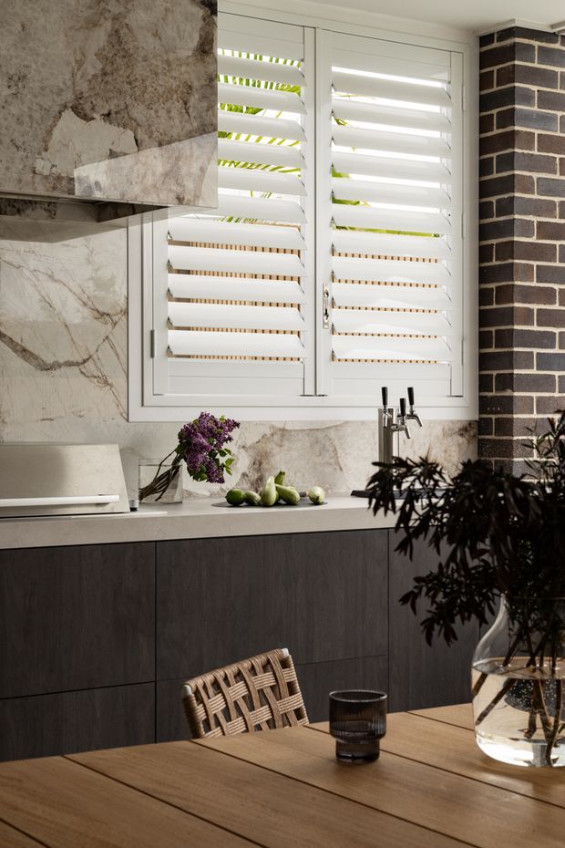 A refined kitchen and dining area featuring dark timber cabinetry, a pale stone benchtop and splashback, a large white plantation-shutter window, and a contrasting dark brick wall. A timber dining table, woven chair, glassware, fruit, and floral arrangement create a warm, sophisticated setting.