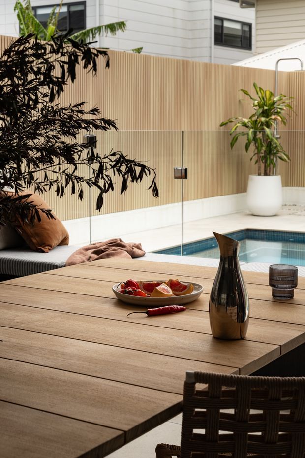 A refined outdoor entertaining area beside a compact swimming pool, viewed across a large timber dining table. The space features a built-in upholstered bench with cushions, woven dining chairs, a tall privacy screen in a vertical timber-slatted finish, potted greenery, and a modern residential building beyond. The palette is warm and natural, with timber, neutral upholstery, white walls, and dark metal accents.