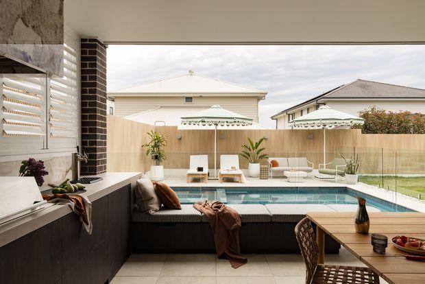 Contemporary covered outdoor entertaining area overlooking a rectangular swimming pool, with a built-in dark kitchen or barbecue counter at left, integrated bench seating, dining table, umbrellas, and a fenced suburban setting beyond. The space uses large pale floor tiles, timber screening, glass pool fencing, and dark brick and joinery accents.