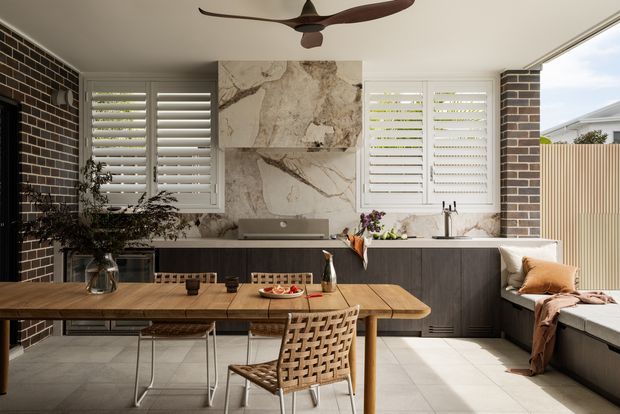 A bright, contemporary kitchen and dining area with a long timber dining table, woven dining chairs, dark cabinetry, a stone-look feature wall and splashback, white plantation shutters, and a built-in window seat. Exposed dark brickwork adds texture, while a ceiling fan and large windows create an airy indoor-outdoor feel.