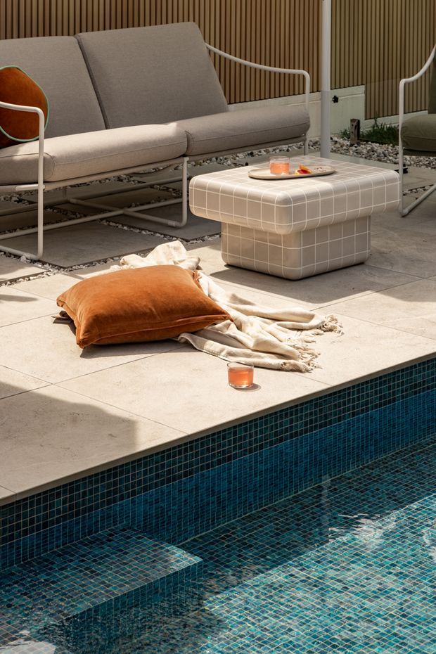 A stylish outdoor poolside lounge area with a deep blue-green tiled swimming pool, pale stone paving, a neutral upholstered metal-framed sofa, a matching rounded ottoman coffee table, and warm terracotta cushions. Vertical timber screening forms the backdrop, while a casual throw and drinks create a relaxed resort-like setting.