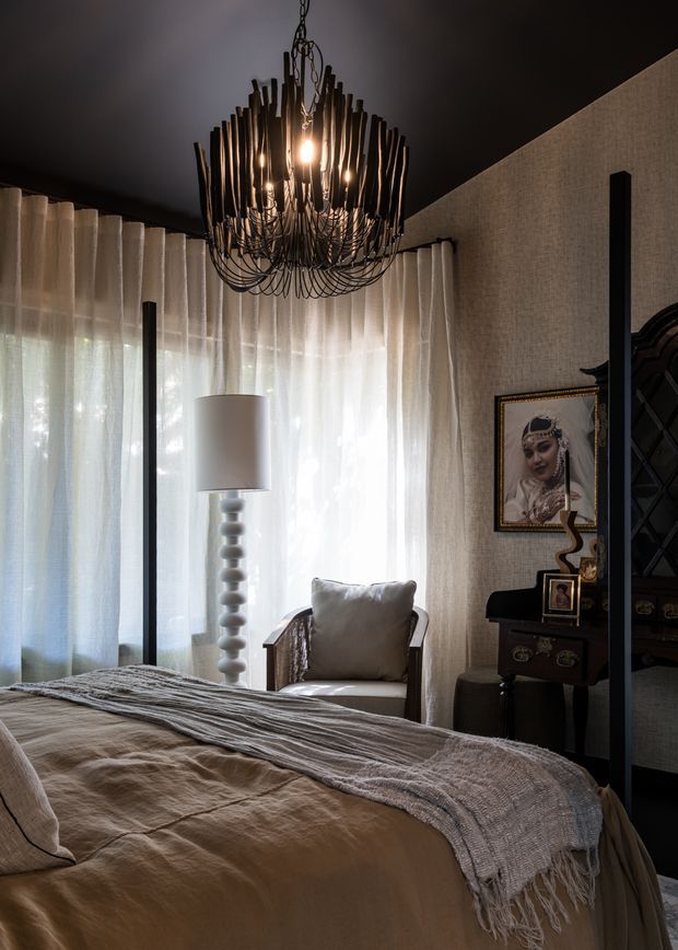 A moody, sophisticated bedroom with a large bed in the foreground, layered neutral bedding, sheer floor-to-ceiling curtains, a sculptural white floor lamp, upholstered armchair, dark side table, framed portrait, and an ornate black branch-like chandelier. The room uses dark walls and ceiling contrasted with soft beige textiles and filtered daylight, creating a luxurious, dramatic atmosphere.