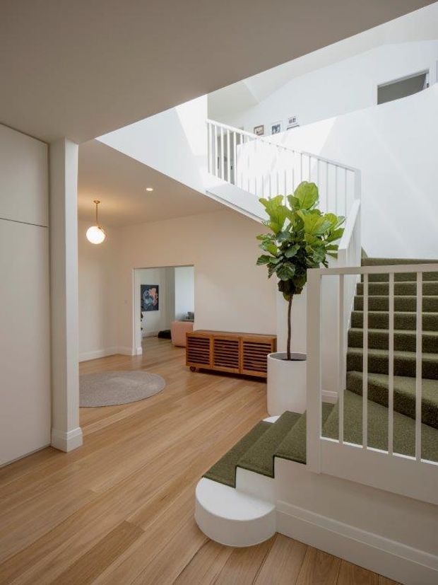 Bright contemporary residential stair hall and entry space with white walls, a white painted balustrade, carpeted staircase, light timber flooring, and an open void above. A sculptural round pendant, potted indoor tree, low timber slatted console, and glimpsed adjoining living area add warmth and visual interest.