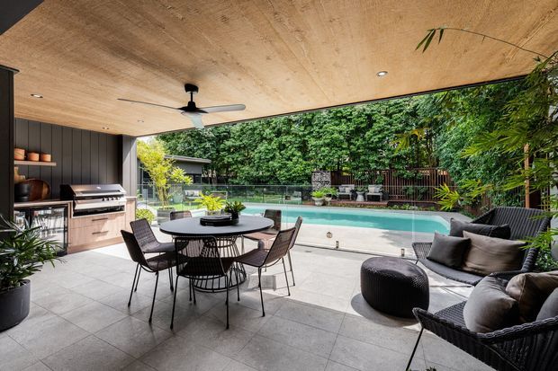 Covered outdoor entertaining area with a tiled patio, built-in outdoor kitchen and barbecue, dining table, lounge seating, ceiling fan, and a swimming pool beyond. Dense greenery and a timber privacy fence frame the relaxed contemporary space.
