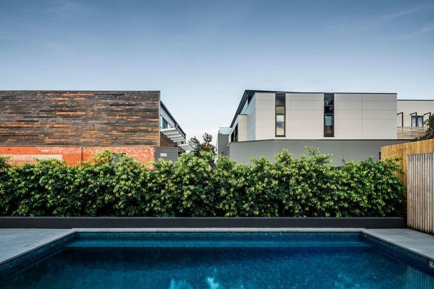 Exterior view of a contemporary residential property with a rectangular swimming pool in the foreground, dense hedge screening, timber fencing, and neighbouring modern homes. The main house features clean geometric lines, pale cladding, dark trim, and narrow vertical windows, while an adjacent structure combines brickwork with weathered timber cladding.