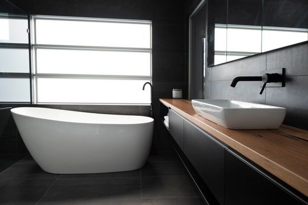 A contemporary dark-toned bathroom featuring a freestanding white bathtub beneath large frosted windows, a floating dark vanity with a warm timber benchtop, white vessel basins, black wall-mounted tapware, and a large mirror. The space has strong contrast between black architectural surfaces, white sanitaryware, and natural timber.