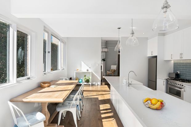 Bright open-plan kitchen and dining area with a long white island, integrated sink, white cabinetry, dark blue-grey tiled splashback, stainless steel range cooker, timber dining table, and large windows bringing in strong natural light. The space has a clean, contemporary feel with pendant glass lighting and dark timber flooring.