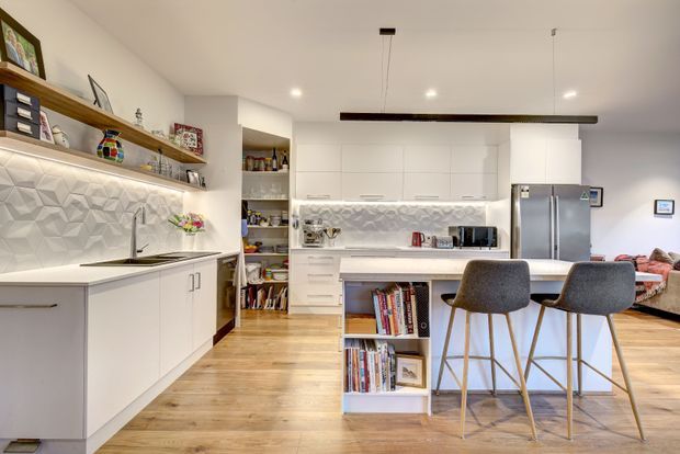 Bright contemporary open-plan kitchen with white flat-panel cabinetry, a central island with integrated open shelving, pale timber flooring, stainless-steel appliances, and a geometric textured splashback. Open shelving and a small pantry area are visible to the left, while a living area is partially visible beyond the kitchen.