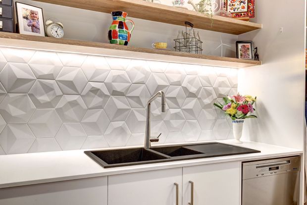 Bright contemporary kitchen or kitchenette wall featuring a double-bowl undermount sink, tall chrome gooseneck tap, white benchtop, white flat-panel cabinetry, integrated dishwasher, and a striking three-dimensional geometric white backsplash. Open timber display shelving above holds decorative objects and framed photographs.