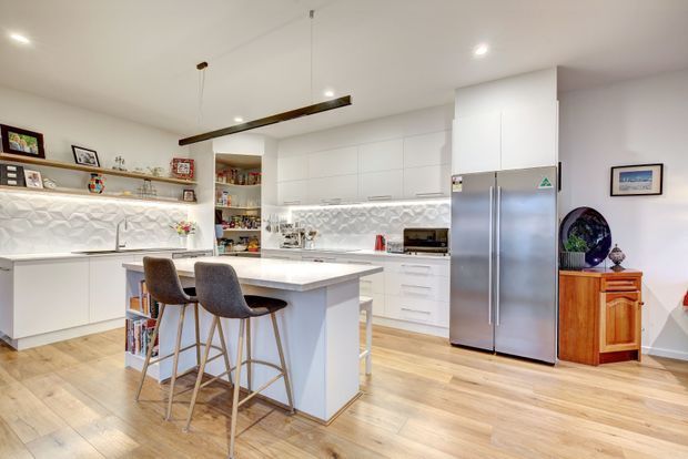 Bright contemporary white kitchen with a central island and two bar stools, extensive white cabinetry, stainless-steel French-door refrigerator, pale timber flooring, and a textured geometric backsplash. Open shelving displays decorative items, while a long linear pendant light hangs above the island. A small timber sideboard and framed artwork sit at the edge of the room.