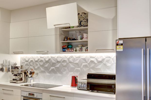 A compact contemporary kitchen with white full-height cabinetry, integrated appliances, a white benchtop, and a faceted geometric white splashback illuminated by under-cabinet lighting. Open shelving displays cups and kitchenware, while a coffee machine, kettle, toaster, cooktop, oven, and large stainless-steel refrigerator occupy the work zone.