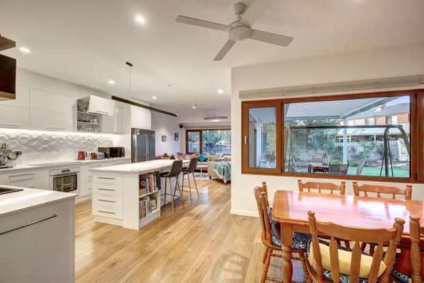 Bright open-plan residential interior showing a contemporary white kitchen with island seating, timber-look flooring, a dining area with a wooden table and chairs, and a living area beyond. Large timber-framed glazing overlooks a garden, while recessed lighting and a ceiling fan contribute to the clean, airy feel.