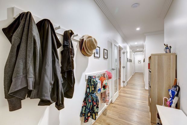 Bright, narrow residential hallway with white walls, decorative ceiling moulding, light timber flooring, and extensive wall-mounted coat and hat storage near the entrance. A tall timber storage cabinet and umbrella stand sit along the right side, while framed artwork and doors line the corridor.