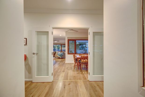 View through an open interior doorway into a bright dining and living area. The space features pale timber flooring, white walls with decorative ceiling moulding, glazed white doors, a timber dining table and chairs, and large windows with reddish timber frames.