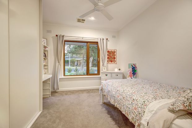 Bright, softly styled bedroom with a large floral-covered bed, beige carpet, white walls and built-in white storage. A timber-framed window with curtains overlooks greenery, while a ceiling fan and small dresser complete the room. The space has a comfortable, light traditional feel with playful artwork and shelving.