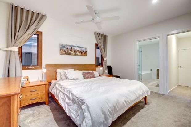 Bright, comfortably furnished bedroom with a large bed, timber headboard and matching bedside furniture, white bedding, carpeted floor, draped windows, ceiling fan and recessed lighting. An open doorway leads directly to a tiled bathroom, with a separate hallway or storage door also visible.