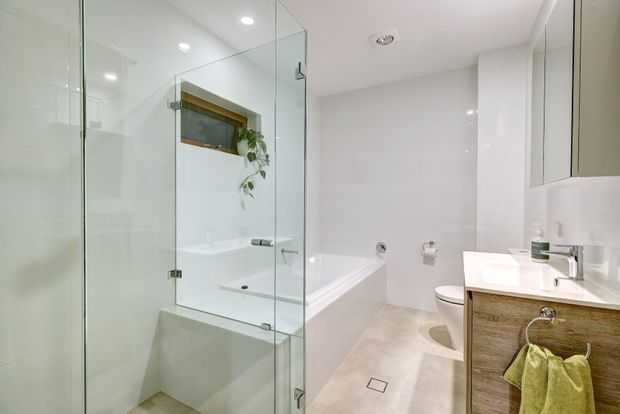 Bright contemporary bathroom with a frameless glass shower enclosure, freestanding bathtub, toilet, wall-mounted mirror cabinetry, and timber vanity with a white basin. The space uses pale tiled surfaces, light flooring, recessed lighting, and restrained natural accents.