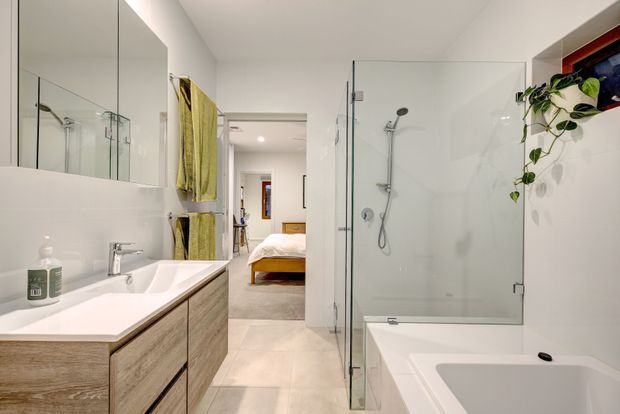Bright contemporary bathroom with a frameless glass walk-in shower, white freestanding-style bathtub, double vanity with light timber cabinetry, large mirrored wall cabinets, and white tiled finishes. A bedroom is visible through the open doorway beyond the bathroom.