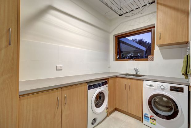 Bright, functional laundry room with L-shaped light timber cabinetry, pale benchtops, white walls, tiled flooring, and front-loading washing and drying appliances. A small timber-framed window provides an exterior view, while a ceiling-mounted drying rack adds practical utility.