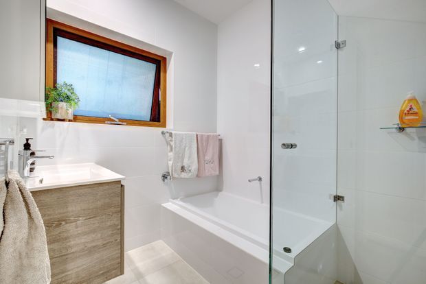 Bright contemporary bathroom with a timber-framed frosted window, white tiled walls, a white bathtub, and a frameless glass shower enclosure. A wood-look vanity with white benchtop, wall-mounted towel rail, and recessed shower shelf complete the clean, functional space.