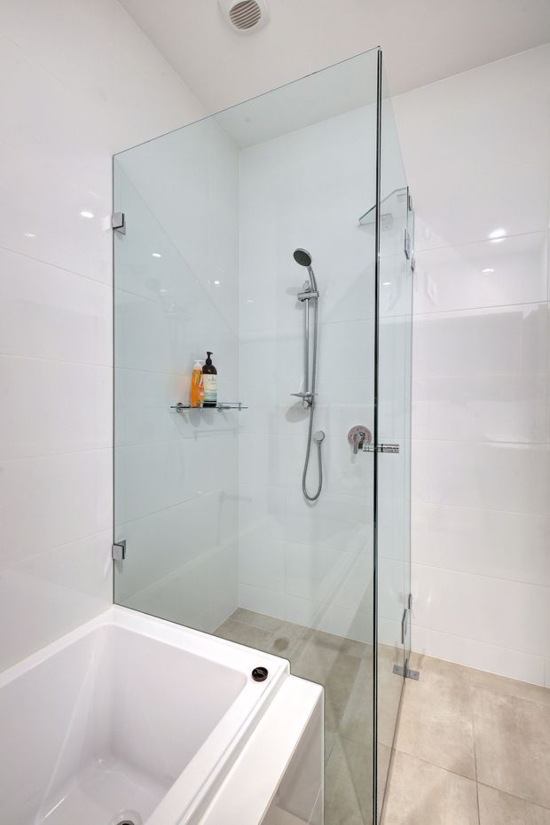 Bright contemporary bathroom featuring a frameless glass walk-in shower with chrome fixtures, a built-in white bathtub, glossy white wall tiles, and large light beige floor tiles.